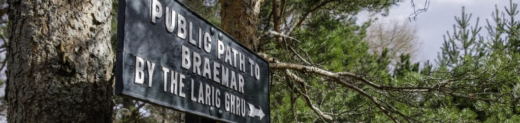 Sun shining on a rectangular sign that says Public Path to Braemar by the Larig Ghru, surrounded by pine trees