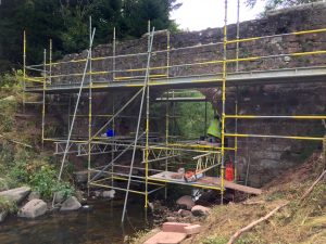 Scaffolding surrounding a stone arched bridge over a burn with a mason working on repairing the mortar.