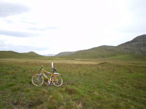 Lochan Dubh, more a bog after a dry spell. The path shown on the map comes through the gap in the hills immediately above the bike, and its line is marked across rather featureless terrain by intermittent wooden posts such as the one used as a prop here. © Richard Law, Geograph