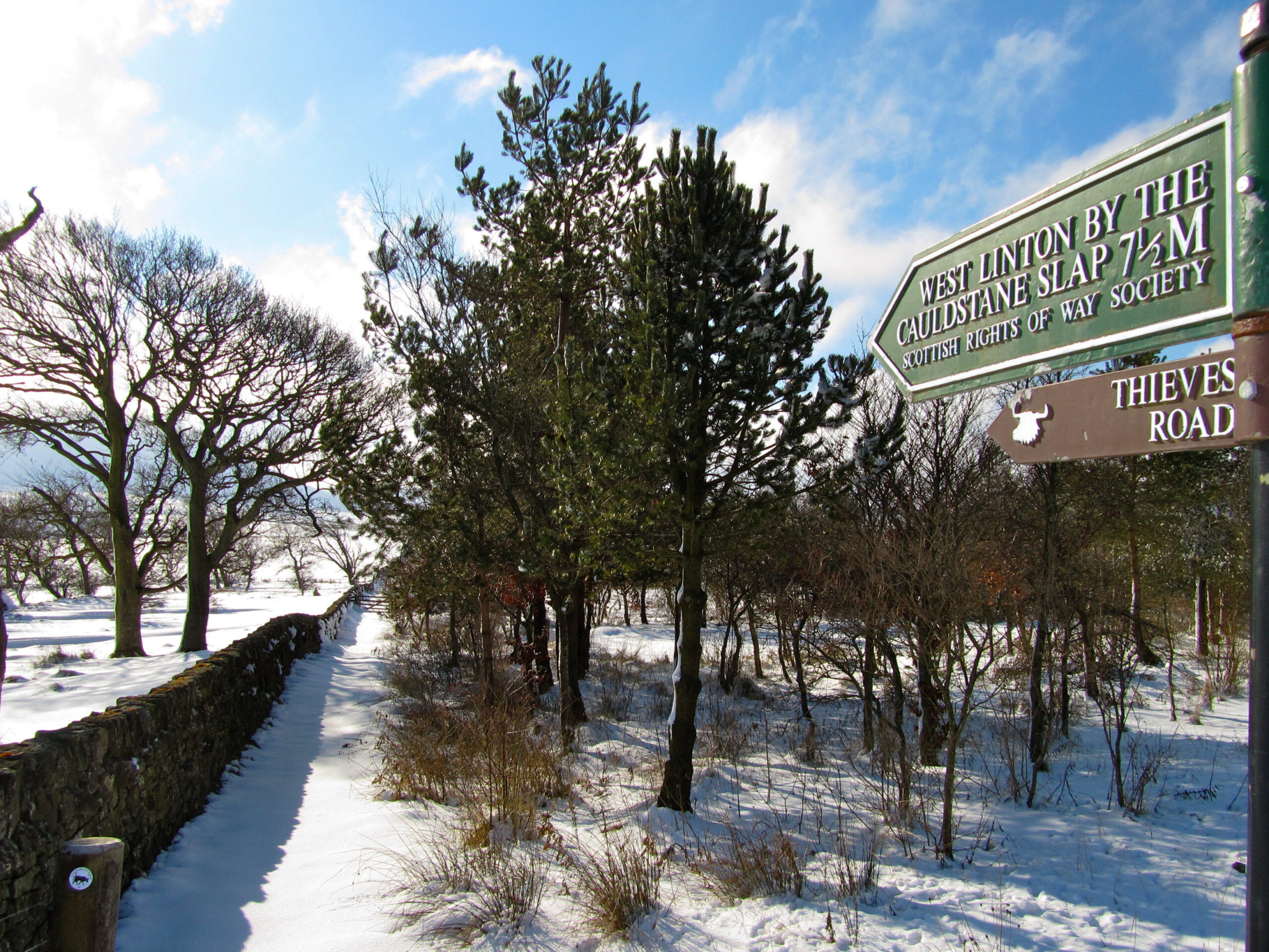 A path between a drystone dyke and a wood. A signpost indicates that the path is called the Thieves Road and a ScotWays sign shows that it is 7.5 miles to West Linton by the Cauldstane Slap.