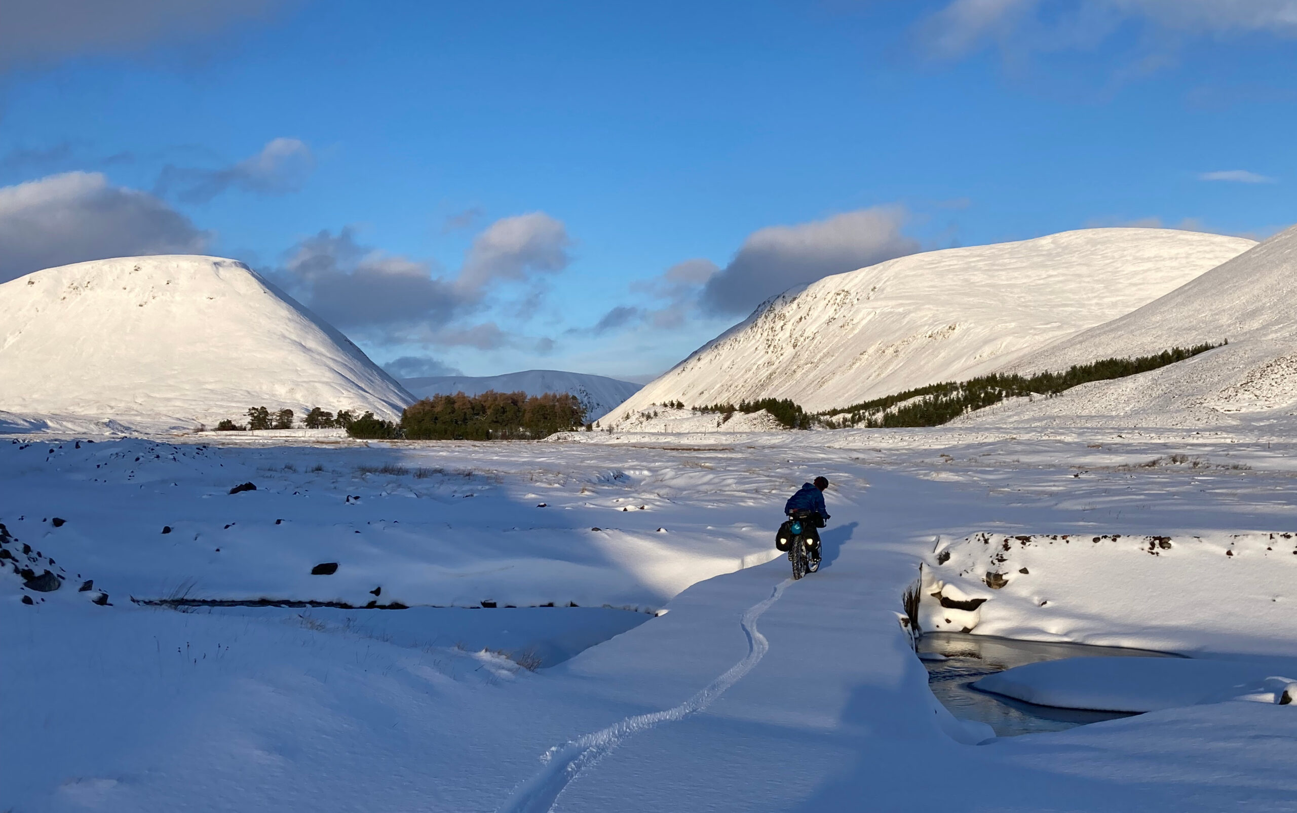 Snow covered hills against a blue sky, a small clump of trees in the distance between the hills. A cyclist leaves the only track in the snow on the bridge across the river.