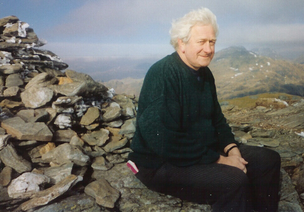 A man with grey hair and a green jumper sits on a cairn, smiling and looking at the camera.
