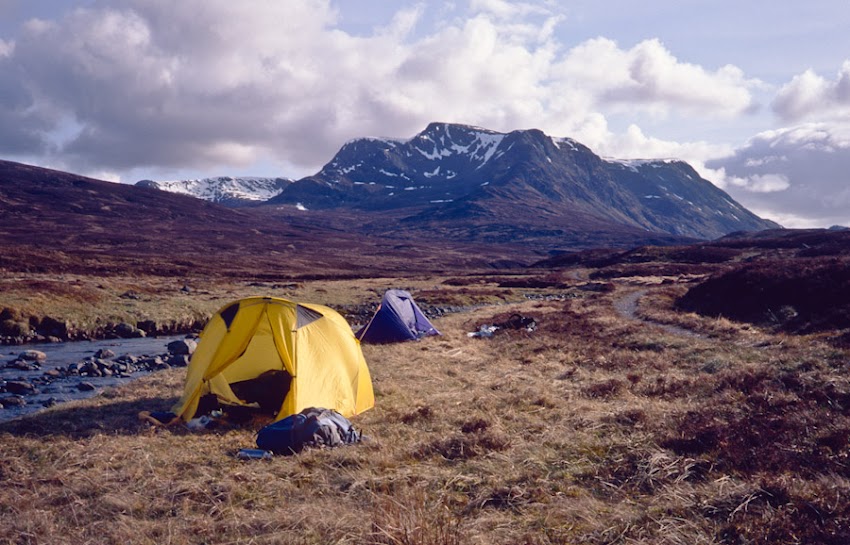A yellow dome tent and a smaller blue tent are pitched on a grassy riverbank in a remote wilderness landscape. A shallow stream flows along the left side of the tents, and the rugged, snow-dusted Ben Alder, rises in the background under a partly cloudy sky.