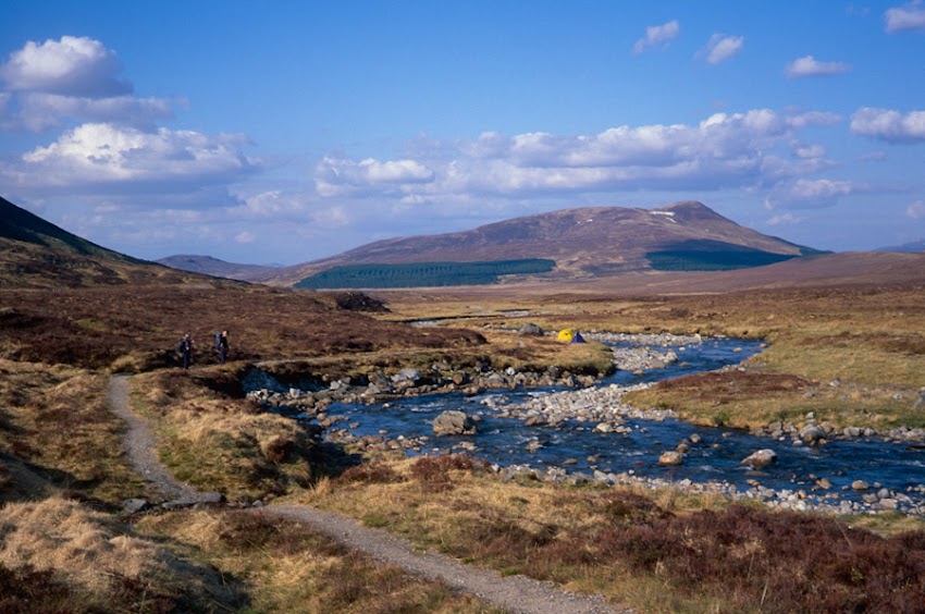 Two hikers walk along a narrow dirt path beside a rocky stream winding through a vast, brown moorland landscape. A yellow and a blue tent are pitched on the riverbank in the mid-distance, with a large, sloping mountain featuring a dark green pine forest line visible under a bright, partly cloudy sky.