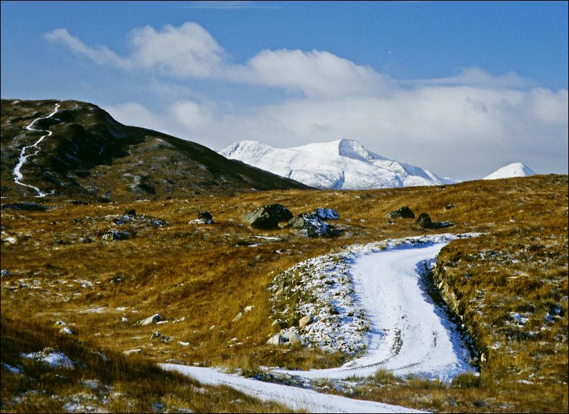 A winding road covered in light snow cuts through a landscape of golden-brown grasses and scattered rocks, leading toward distant, snow-capped mountains under a bright blue sky with wispy clouds.