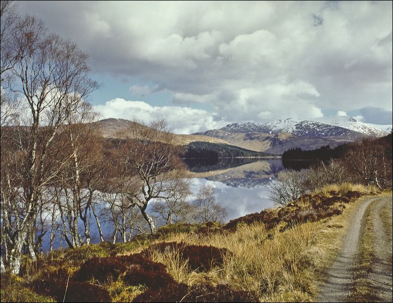 A gravel path winds along the shore of Loch Ossian. The calm, reflecting lake is surrounded by moorland grass and bare deciduous trees, with snow-dusted mountains rising in the distance under a partly cloudy sky.
