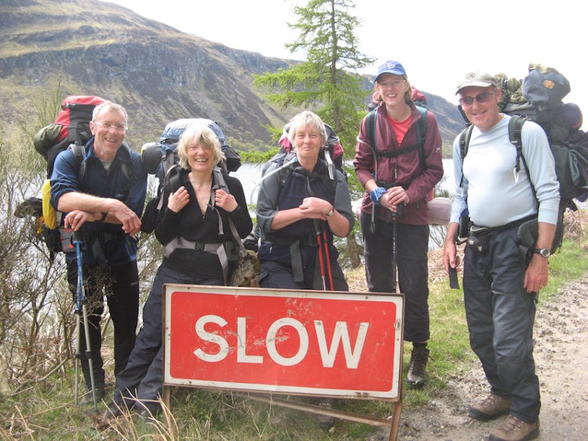 Five hikers carrying large backpacks pose together behind a red rectangular "SLOW" sign on a mountainous trail, with a loch and hilly landscape visible in the background.