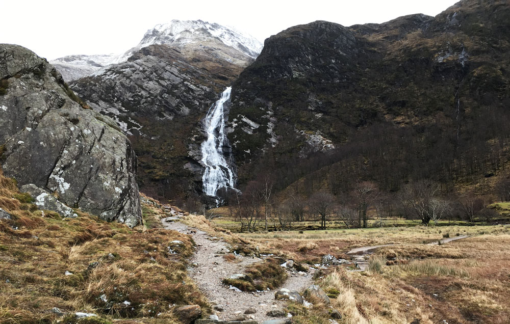 A rugged path leads through a dry, grassy glen toward a tall waterfall cascading down a rugged, dark mountainside with a snow-capped peak in the distance.