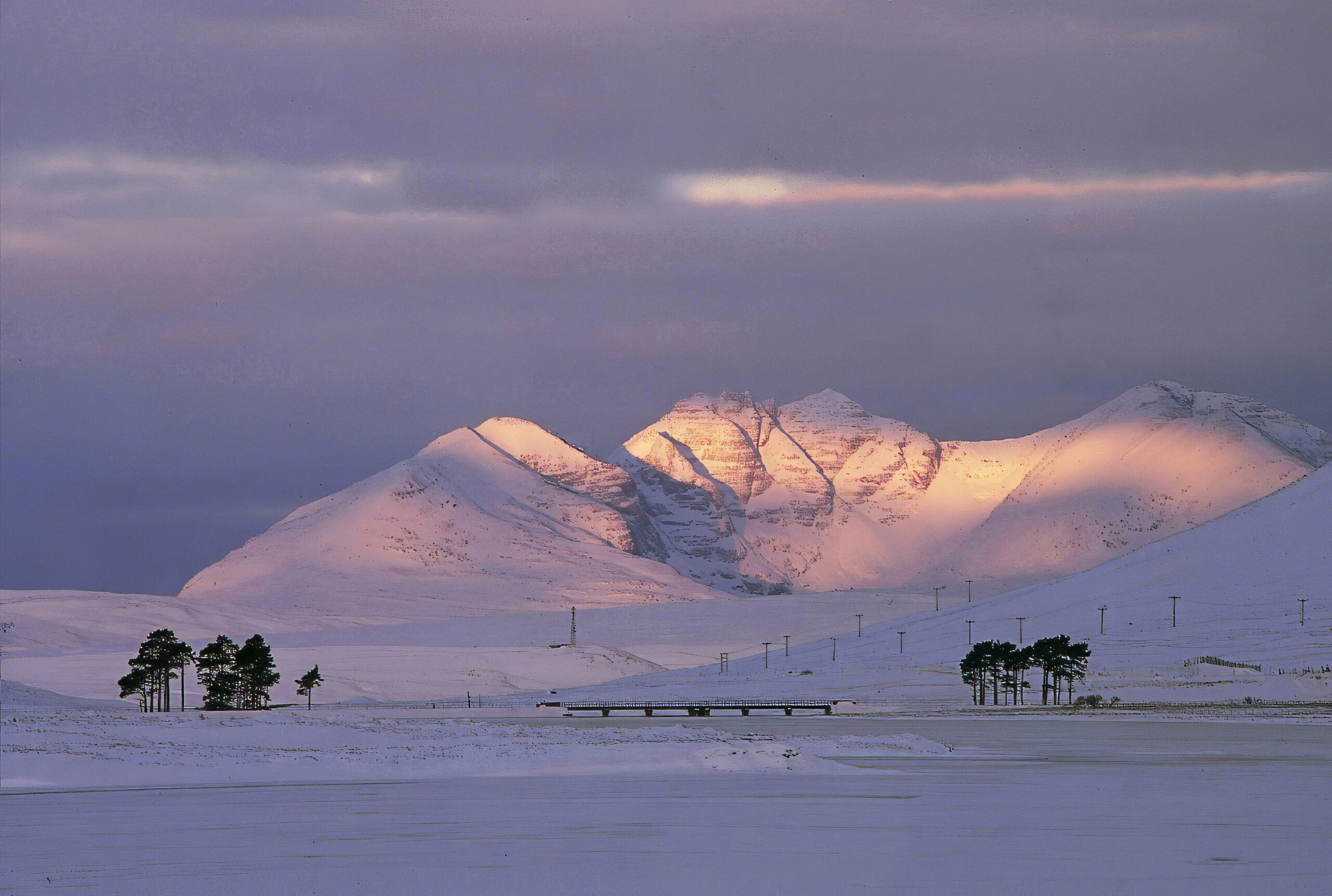 A snowy mountain illuminated by a pink sunset. There is a small bridge and trees underneath the mountain.