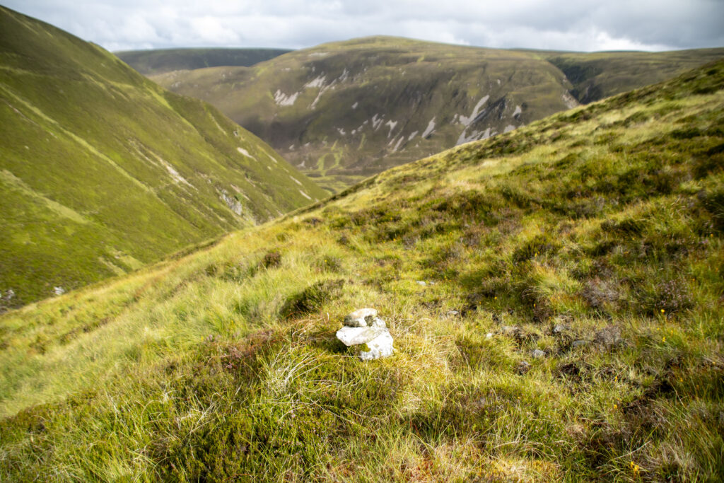 A small stone cairn sits on a grassy, heather-covered slope overlooking a vast, rolling mountain valley beneath a cloudy sky.